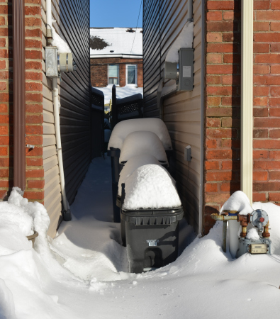 a row of trash bins covered with snow, between two brick houses