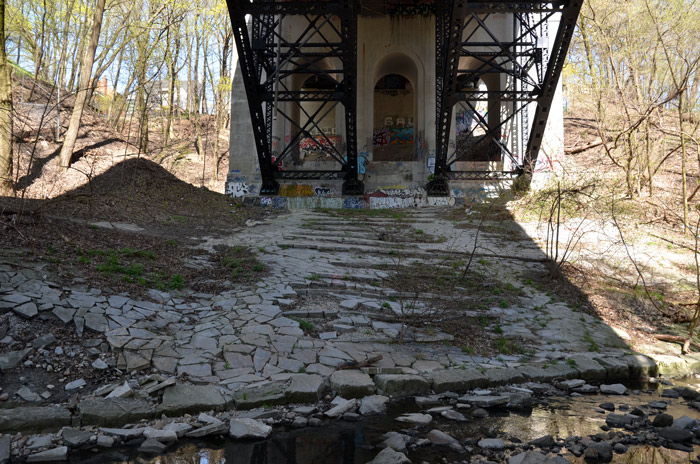 under a concrete and steel bridge.  There is a creek in the foreground.  Lots of small trees are in the picture.  It is early spring and the leaves on the trees are just starting to show. There was once a concrete floor like structure under the bridge but is very cracked and broken.  There is graffiti on the bridge.