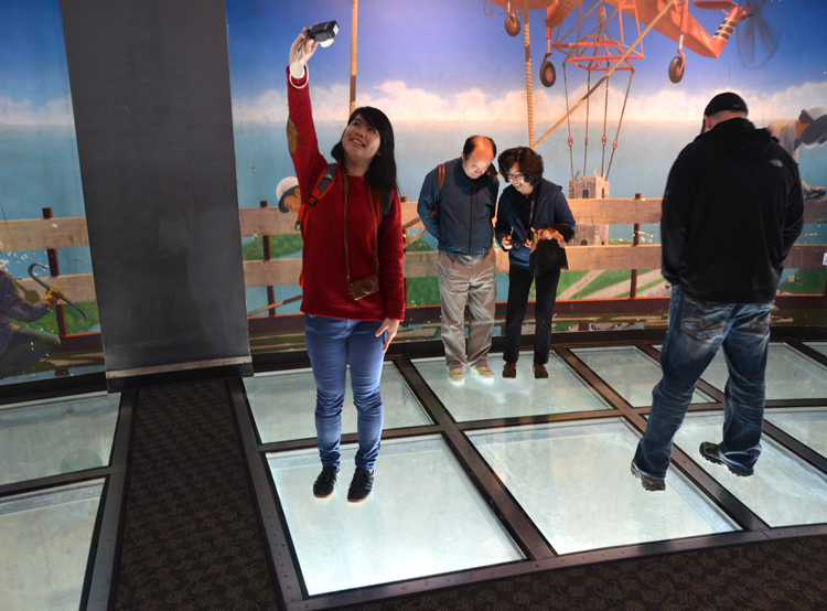 people are looking down through the glass floor on the observation deck of the CN Tower.  Others are taking selfies there.  