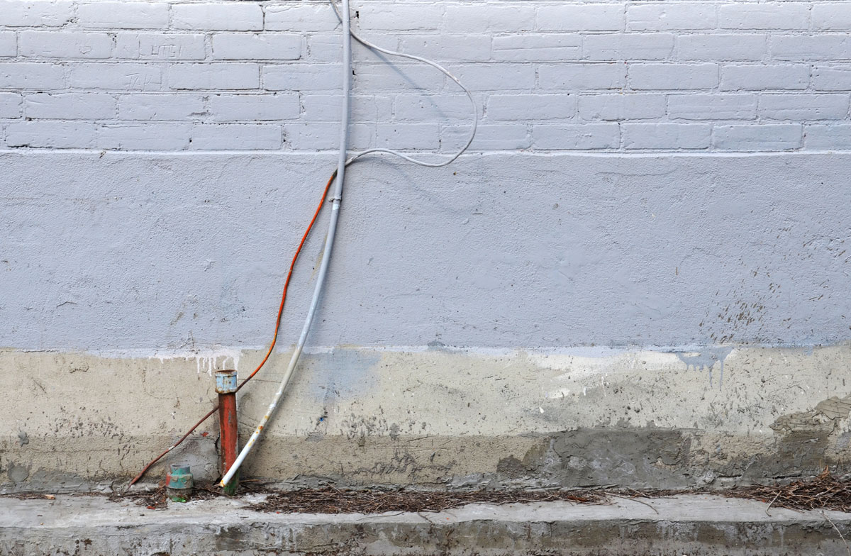 a short copper pipe sticks out of the kerb beside a pale grey wall.  Two wires run down the side of the wall towards the pipe, one metal and one painted the same colour as the wall.   