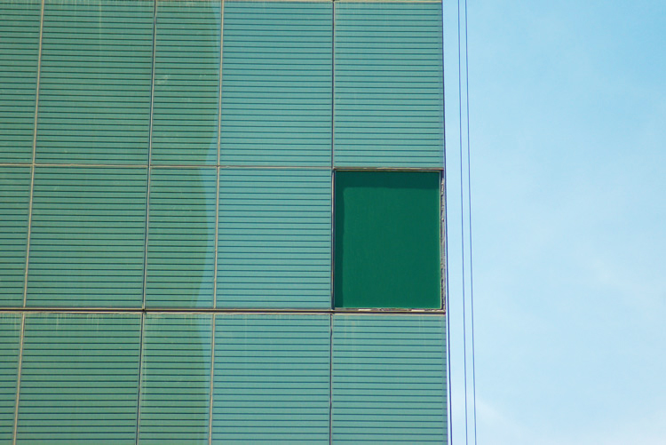 abstract looking picture of wall of a new building that is two tones of green, with a rectangular shape ina darker green.  Light blue sky beside the wall.  