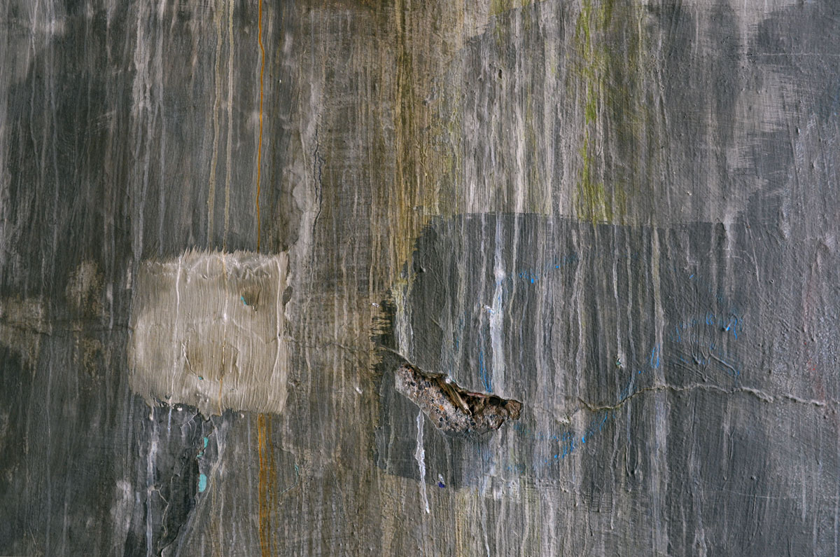 abstract looking picture of an old concrete wall that has started to crack in some places.  Part of the wall was patched with a bit of fabric like substance and it is fraying a bit. 