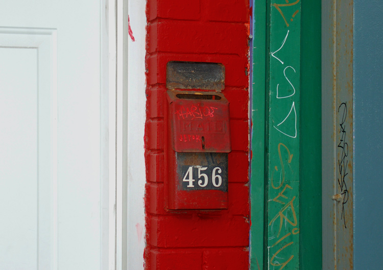 abstract looking picture of a wall beside  a white door.  There is a small section of brick painted bright red, and then some green wood.  The mailbox is on the brick and it too is painted bright red.  The number on the box is 456.  Graffiti tag LSD on the green. 