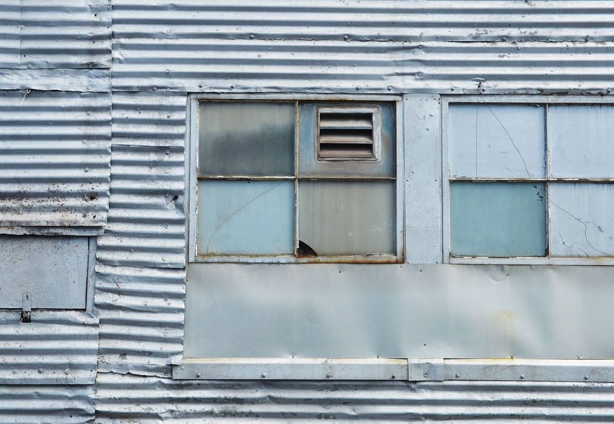 abstract looking picture of an old corrugated metal wall.  what used to be a large window has been covered with metal, and a few small windows have been either painted over or covered from the inside