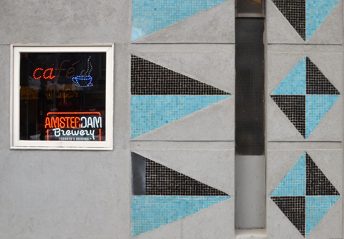 abstract looking picture of part of the exterior wall of the Rex Hotel.  Triangle and diamond shapes are made of turquoise and black tiles, set into grey concrete.  A square window is also  in the picture, with ights that say cafe as well as Amsterdam Brewery.  The cafe sign is half burned out. 