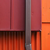 abstract looking picture of the intersection of an orange wood wall and a maroon metal wall.  A brown downpipe runs along the outside of the maroon part.   A greenish shrub grows in front of the orange part.  