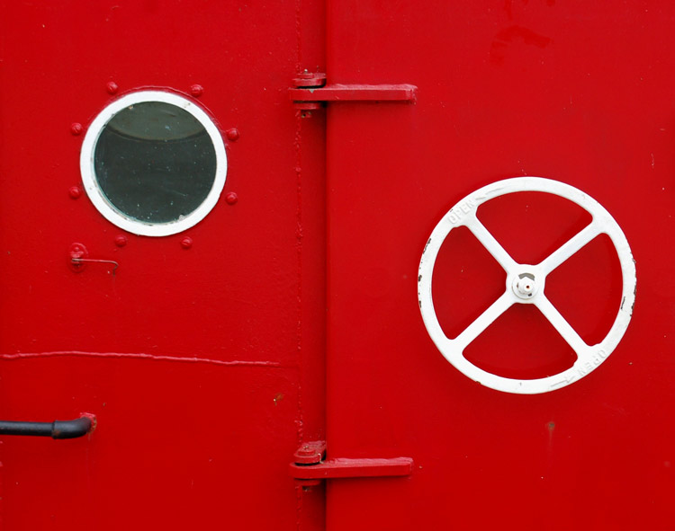 abstract looking picture of the exterior wall of bright red tug boat with a small circular window with a white frame and a white steering wheel.  
