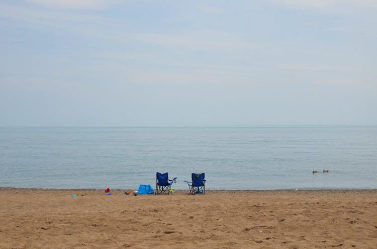 two empty beach chairs on the beach.  Photo is taken from behind the chairs and looking towards the lake 