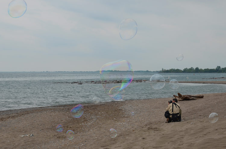 A man crouching down on the beach to get a better angle as he photographs large bubbles that are floating in the breeze. 