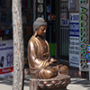 a large brown statue of a seated buddha is on the sidewalk outside a store.  A bike is parked near it and a woman is walking past. 