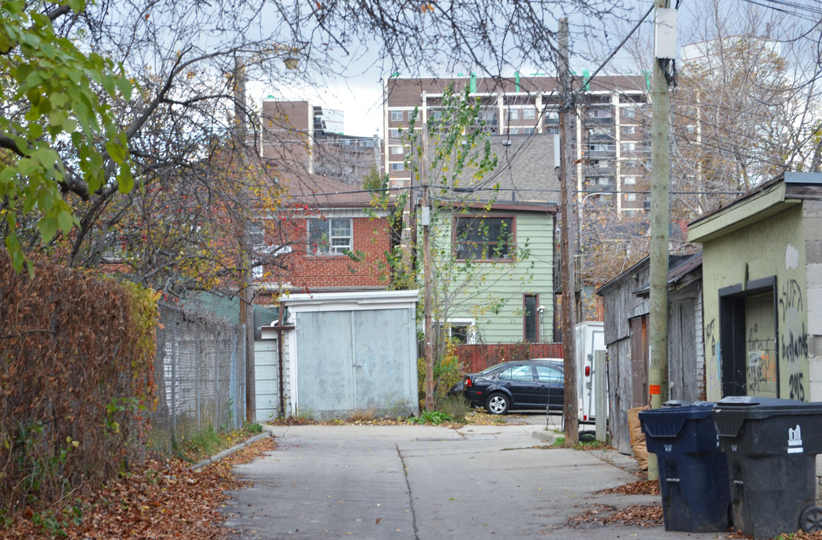 a Toronto alley, garbage bins, garages, the back of two houses, one very crooked, the back of a low rise apartment building, chainlink fence, fall foilage. 