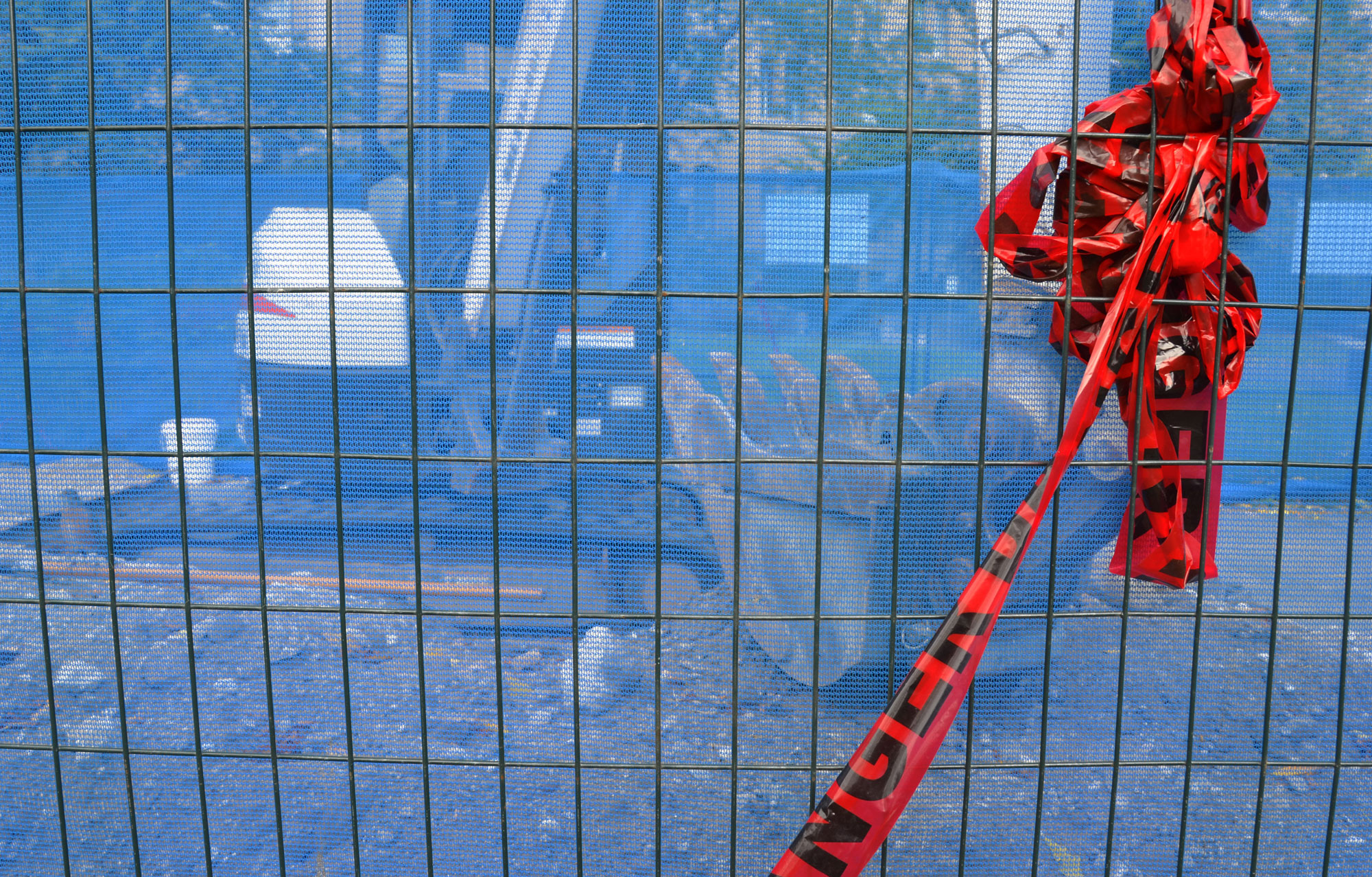 a very blue fence around a construction site.  You can just see a digger through the fence.  Some red plastic ribbon is hanging from the top of the fence. 