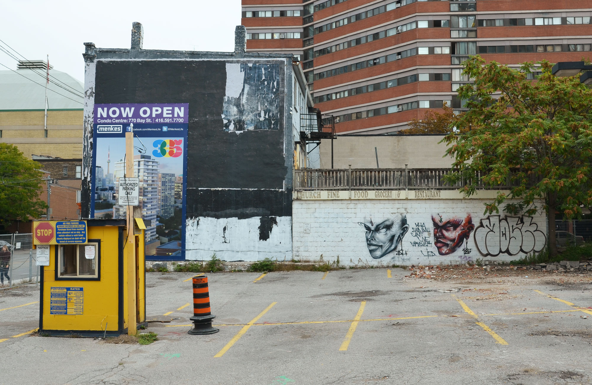 an empty parking lot with an old yellow hut for the parking attendant, the far wall of the lot is concrete and has been painted with two graffiti faces