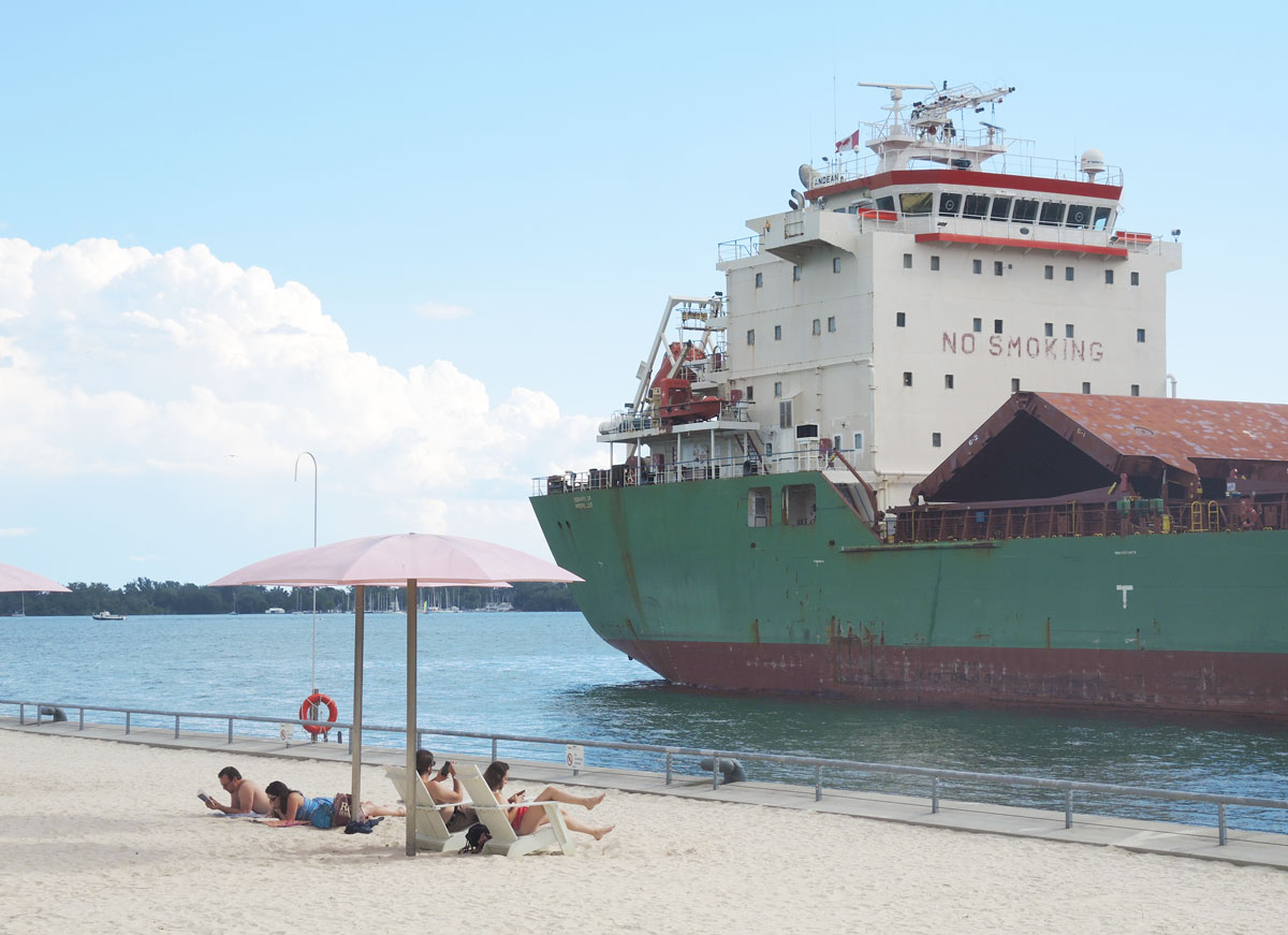 a large ship, or lake freighter, is parked in the harbour beside sugar beach where people are sitting under pink umbrellas.