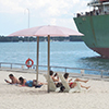 a large ship, or lake freighter, is parked in the harbour beside sugar beachwhere people are sitting under pink umbrellas.