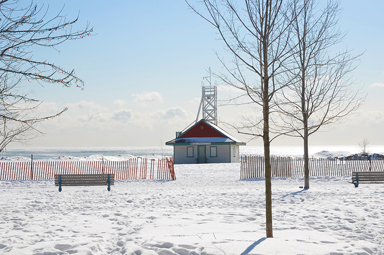 Leuty Lifesaving station, a small white wood building with a red roof, in the winter, surrounded by snow.  A few trees and empty benches are in the picture. 
