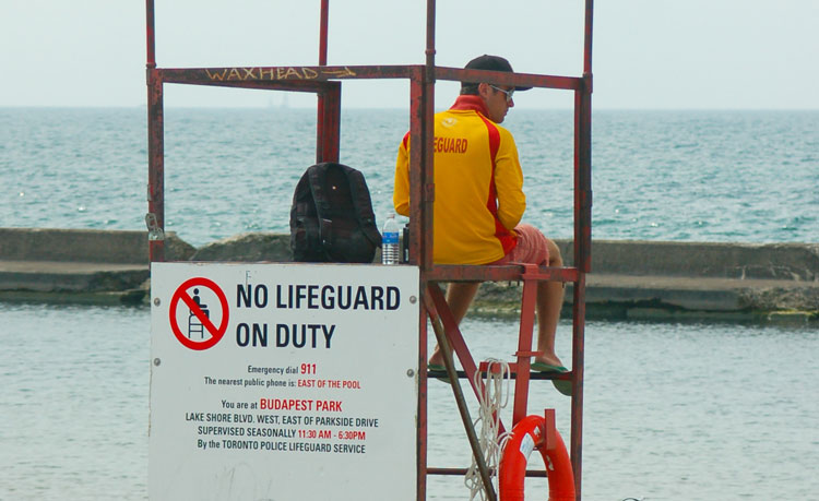 a lifeguard in yellow jacket is sitting on a lifeguard chair at Bucharest Park even though the sign behind him says 'No lifeguard on duty'.  