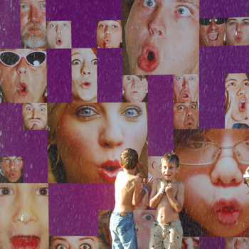 A boy playing in a fountain.  The fountain is a very large tower shape and it is covered with many faces.  The water comes out of the mouths of the faces. 