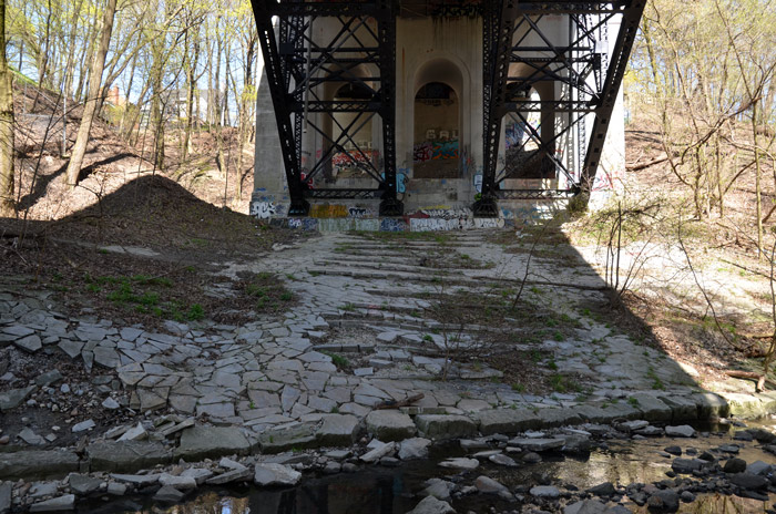 under a concrete and steel bridge. There is a creek in the foreground. Lots of small trees are in the picture. It is early spring and the leaves on the trees are just starting to show. There was once a concrete floor like structure under the bridge but is very cracked and broken. There is graffiti on the bridge.