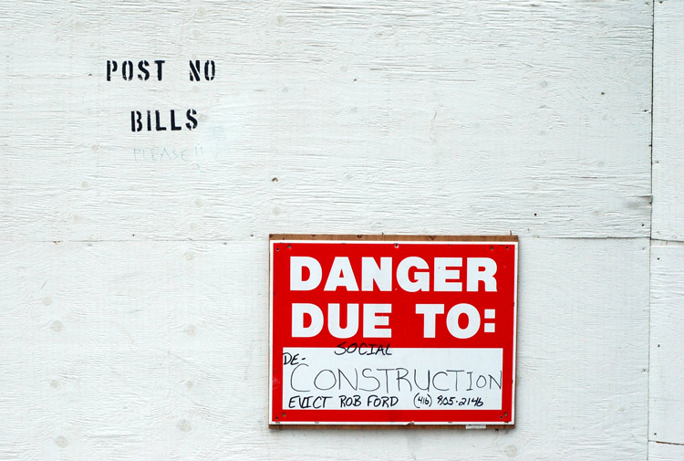 on a white fence that says post no bills, there is a red and white danger sign that now reads danger due to social deconstruction