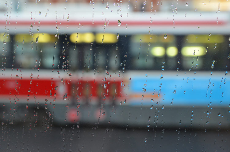 looking through one streetcar window on a rainy day towards the side of another streetcar.  Water drops on the window. 