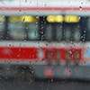 looking through one streetcar window on a rainy day towards the side of another streetcar.  Water drops on the window. 