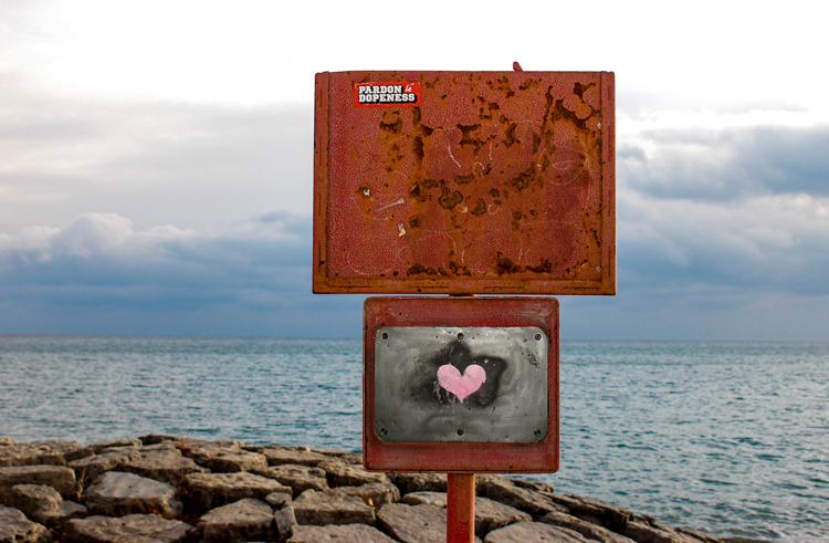 a rusty old sign on the waterfront that someone has painted a pink heart on.  Lake Ontario is in the background 