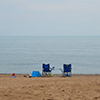 two empty beach chairs on the beach.  Photo is taken from behind the chairs and looking towards the lake 