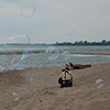 A man crouching down on the beach to get a better angle as he photographs large bubbles that are floating in the breeze. 