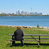 A man sits at a picnic table in a park overlooking Lake Ontario.  The Toronto skyline and the CN are in the distance 