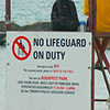 a lifeguard in yellow jacket is sitting on a lifeguard chair at Bucharest Park even though the sign behind him says 'No lifeguard on duty'.  